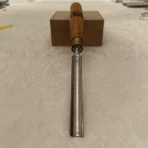 May include: A woodworking chisel with a wooden handle and a silver-colored blade. The handle is a light brown color with a brass ferrule. The chisel is resting on a small, light brown wooden block.