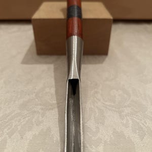 May include: A woodworking gouge featuring a long, polished metal blade with a U-shaped profile and a wooden handle. The handle is reddish-brown with a dark band near the center and a metallic cap at the top end. The tool is positioned vertically.