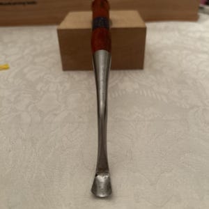 Puede incluir: Una gubia para madera con una hoja de metal curva de acabado pulido y un mango de madera largo y cilíndrico. La herramienta tiene una virola de metal distintiva que conecta el eje de acero con la empuñadura de madera de color marrón rojizo.