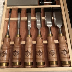 May include: A set of six Lorentz Fine Tools wood chisels with light wood handles, brass ferrules, and steel blades, arranged in a wooden storage box. The set includes a rectangular sharpening stone and a black metal honing guide tool.