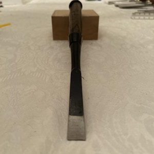 May include: A woodworking chisel with a dark wooden handle and a black metal shaft. The blade is silver and rectangular. The chisel is resting on a small, light brown wooden block.