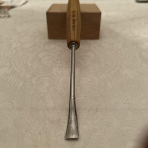 May include: A metal wood carving gouge with a long, slender, curved steel blade and a light brown wooden handle. The handle features the text SWISS MADE 25/10 printed clearly on the side. The tool is positioned vertically with the blade pointing down.