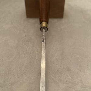 May include: A woodworking chisel with a wooden handle and a metal blade. The handle is a dark brown color, and the blade is silver. The chisel is positioned vertically against a neutral background.