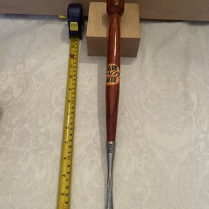 May include: A long, slender wood carving gouge with a polished dark brown wooden handle and a metal blade. A yellow measuring tape placed alongside shows the tool is approximately 13.5 inches long. A circular sticker with Japanese text is on the handle.