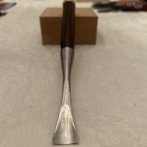 以下が含まれることがあります： 湾曲した研磨済みの金属製ブレードと円筒形のダークブラウンの木製ハンドルを備えた木彫り用彫刻刀。このツールは、細長い金属製のシャフトを持ち、木材を削るための幅広で凹状の刃先へとつながっています。