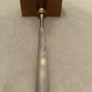 May include: A woodworking chisel with a dark wooden handle and a silver blade. The blade has the text "Sheffield Marples England" etched on it. The chisel is resting on a wooden block.