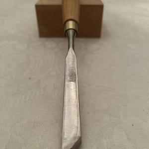May include: A wood chisel with a wooden handle and a metal blade. The blade has a beveled edge and the brand name "GREENLEE ENGLAND" is visible. The chisel is positioned in front of a small wooden block.