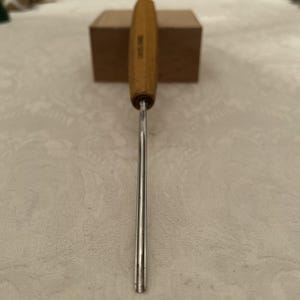 Puede incluir: Una herramienta para carpintería con un mango de madera marrón claro con la inscripción "SWISS MADE" y un vástago de metal plateado. El vástago termina en punta. Herramienta de precisión.