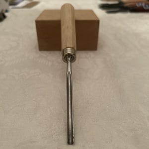 May include: A wood carving gouge with a light-colored wooden handle and a silver-colored metal blade. The gouge is positioned vertically, with a brown block in the background.