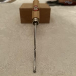 Puede incluir: Un formón para carpintería con mango de madera y hoja de metal. El mango es de madera clara con un emblema rojo y blanco. La hoja es plateada y se extiende hacia abajo. Un bloque de madera está en el fondo.