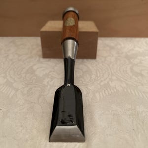 以下が含まれることがあります： 暗く磨かれた仕上げの幅広で平らな鋼鉄製の刃と、面取りされた刃先を持つ日本式の木工用ノミ。ハンドルは明るい茶色の木材で作られており、金属製の口金と上部には円形の金色のインレイが施されています。
