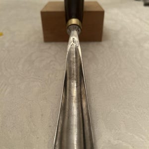 Könnte beinhalten: Ein Holzbeitel aus Metall mit einer langen, gebogenen Stahlklinge mit polierter Oberfläche und einem dunkelbraunen Holzgriff. Das Werkzeug verfügt über eine messingfarbene Metallzwinge, die den Griff mit der Klinge verbindet, welche kleine Gravuren aufweist.