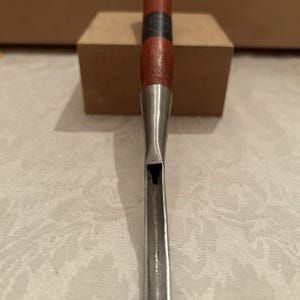 May include: A wood carving gouge featuring a long, curved metal blade and a cylindrical wooden handle. The handle is stained a reddish-brown color with a dark band near the center and the word "KOPPENTZ" stamped clearly near the top end.