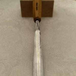 May include: A wood carving gouge featuring a long, curved metal blade with a polished finish and a light brown, octagonal wooden handle. The metal shaft is stamped with Japanese characters near the base of the handle, which is also marked with text.