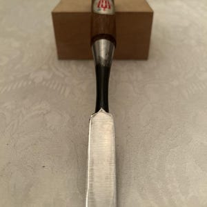 May include: A wood chisel with a dark brown wooden handle and a metallic ferrule. The handle features a small white label with a red emblem and black text. The blade is made of polished steel, transitioning from a dark neck to a flat, sharp edge.