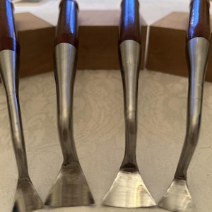 May include: Four metal and wood tools with curved, silver blades and brown wooden handles. The tools are arranged in a row, with the blades facing forward. The handles have a shiny, polished finish.