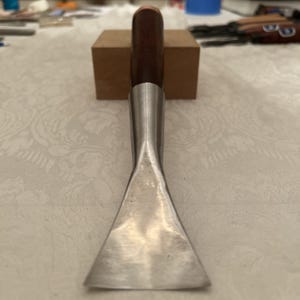 May include: A metal and wood leatherworking tool with a polished silver blade and a dark brown wooden handle. The handle has a copper-colored end. The tool is resting on a small, light brown wooden block.