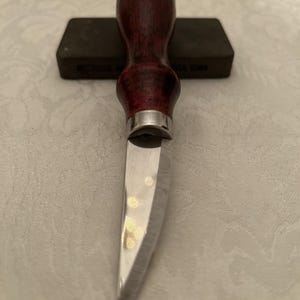 May include: A close-up of a knife with a dark red wooden handle and a silver bolster. The blade is shiny and reflective. A dark gray sharpening stone is in the background.