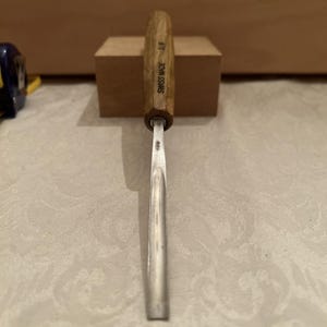 May include: A wood carving gouge with a light brown wooden handle and a curved metal blade. The handle is stamped with the text SWISS MADE and the number 5/8. The metal shaft features a small engraved mark near the base of the wooden handle.