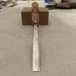 Puede incluir: Un formón para carpintería con mango de madera y hoja de metal. El mango es de color marrón claro con una etiqueta roja y blanca. La hoja es plateada y tiene grabado el número 1. El formón está sobre un bloque de madera.