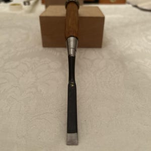 May include: A Japanese woodworking chisel featuring a long, dark metal blade with a sharp, polished edge and a light brown wooden handle. The tool has a metal ferrule connecting the handle to the blade, which displays small, gold-colored markings.