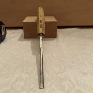 May include: A metal wood carving gouge with a light brown wooden handle. The handle is stamped with the text SWISS MADE and 7/8. The metal blade has a curved, U-shaped profile and features a small stamped logo near the base of the handle.
