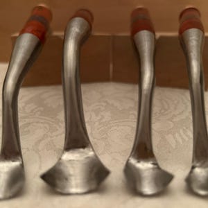May include: A set of four metal spoons with curved handles and amber-colored translucent accents at the top. The spoons feature a polished silver-tone finish and a distinctively shaped bowl, arranged in a row on a light-colored surface.