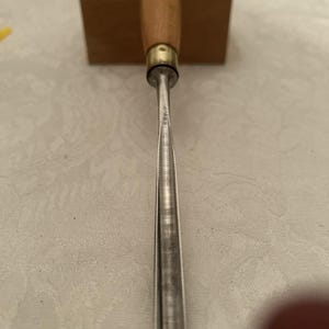 May include: A wood chisel featuring a wooden handle and a metal blade. The handle is light brown with a brass ferrule. The blade is silver and tapers to a point. A wooden block is in the background.
