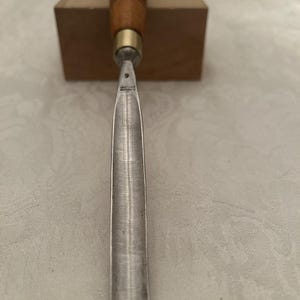 May include: A woodworking chisel with a wooden handle and a metal blade. The handle is brown with green and gold accents. The blade is silver and has the number "9" stamped on it. The chisel is set against a wooden block.