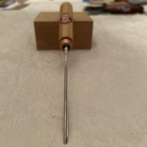 Puede incluir: Una herramienta para cigarros con mango de madera y hoja de metal. El mango tiene una banda color cobre y una etiqueta roja y dorada. La herramienta está colocada sobre un bloque de madera.