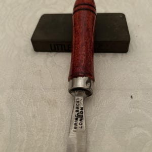 Può includere: Uno scalpello da falegname con un manico in legno rosso scuro e una lama in metallo argentato. La lama reca l'incisione "ERRING BROS LONDON". Sullo sfondo una pietra per affilare grigio scuro.