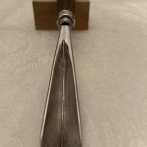 Puede incluir: Primer plano de un formón para carpintería. El formón tiene un mango de madera oscura, una férula de metal plateado y una hoja afilada y angulada. La hoja está colocada contra un bloque de madera.