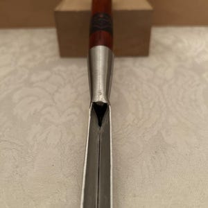 May include: A wood carving gouge featuring a long, polished metal blade with a V-shaped profile and a dark brown wooden handle. The handle includes a copper-colored end cap and a black label with white text near the metal ferrule.