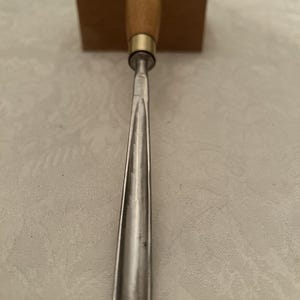 May include: A wood chisel featuring a light brown wooden handle with a brass ferrule and a silver metal blade. The blade tapers to a point. A wooden block is positioned behind the chisel.