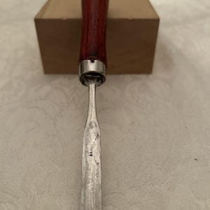 Può includere: Uno scalpello da legno con un manico in legno rosso scuro e una ghiera in metallo argentato. La lama è argentata e reca la scritta "B&GLI". Lo scalpello è posizionato davanti a una scatola di cartone.