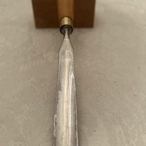 May include: A woodworking chisel with a wooden handle and a metal blade. The handle is light brown with a brass ferrule. The blade is silver and tapers to a sharp edge. The chisel is designed for shaping wood.
