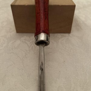 May include: A woodworking chisel with a dark red wooden handle and a silver metal ferrule. The blade is silver and has the number 9 etched on it. A brown cardboard box is in the background.