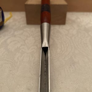 May include: A wood carving gouge featuring a long, curved metal blade with a sharp, U-shaped cutting edge. The tool has a cylindrical wooden handle with a dark brown finish and a metal ferrule connecting the handle to the steel shaft.