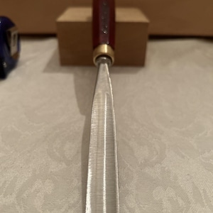 May include: A wood carving gouge featuring a long, curved metal blade with a polished finish. The tool has a cylindrical, dark reddish-brown wooden handle connected to the blade by a circular brass ferrule. The metal surface shows fine texture.