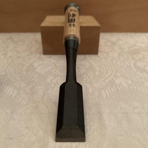 May include: A Japanese wood chisel featuring a wide, flat, dark gray metal blade and a light-colored wooden handle. The handle displays black Japanese kanji characters printed vertically near the top, with a metal ferrule at the base of the grip.