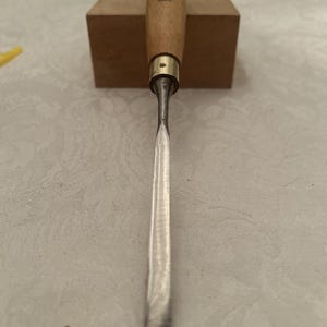 May include: A wood chisel featuring a polished steel blade and a wooden handle. The handle has a brass ferrule and the brand name "GREENLEE" is visible. The chisel is designed for woodworking and carving.
