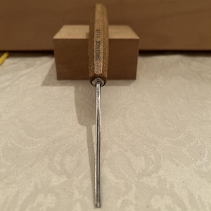 May include: A wood carving gouge with a light brown wooden handle and a metal shaft. The handle is stamped with the text SWISS MADE 11/2, and the metal shaft features a small engraved logo. The tool has a narrow, curved metal blade tip.