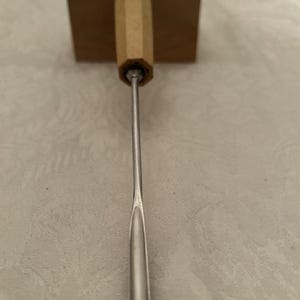 Puede incluir: Una herramienta de metal con mango de madera. El mango es de color marrón claro y facetado. La herramienta tiene una hoja larga y delgada de color plateado. Parece ser un instrumento especializado, posiblemente para artesanía o carpintería.