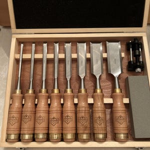 May include: A set of eight wood chisels with wooden handles and metal blades, each marked with sizes from 6mm to 38mm. The set includes a black metal honing guide and a rectangular gray sharpening stone, all organized in a wooden storage case.