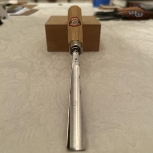 Puede incluir: Un formón para carpintería con mango de madera y hoja de metal. El mango tiene un emblema rojo y dorado. La hoja es plateada y termina en punta. El formón está sobre un bloque de madera.