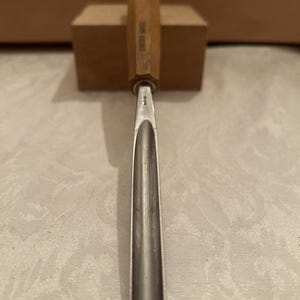 May include: A wood carving gouge with a long, curved metal blade and a light brown wooden handle. The handle features the text "SWISS MADE" stamped into the wood. The metal blade has a deep, U-shaped profile and a sharp, rounded tip.