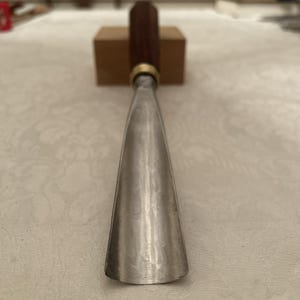 Puede incluir: Un formón para carpintería con una hoja de acero pulido y un mango de madera oscura. El mango es cilíndrico y está unido a la hoja con una virola de latón. El formón está diseñado para dar forma y tallar madera.