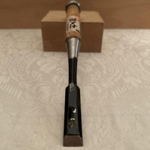 May include: A Japanese wood chisel featuring a light-colored wooden handle with a metal hoop at the top and black Japanese kanji characters printed on the wood. The metal shaft transitions into a flat, black-coated blade with a sharp steel edge.