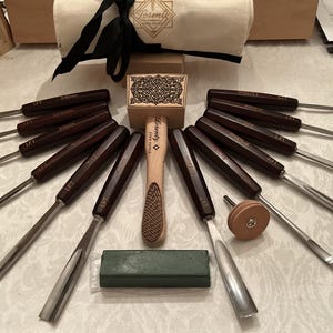 May include: A set of twelve wood carving chisels with dark brown wooden handles and metal blades, a wooden mallet with a decorative top, a small circular sharpening tool, and a rectangular green sharpening block, all branded with Lorentz Fine Tools.