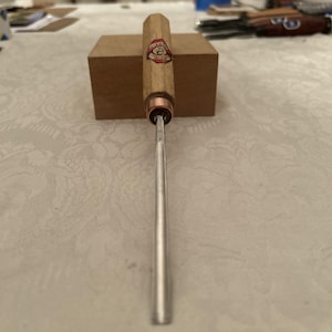 Puede incluir: Una herramienta para tallar madera con un mango cilíndrico de madera y una hoja de metal. El mango tiene una férula de cobre y una etiqueta. La herramienta descansa sobre un pequeño bloque de madera rectangular.